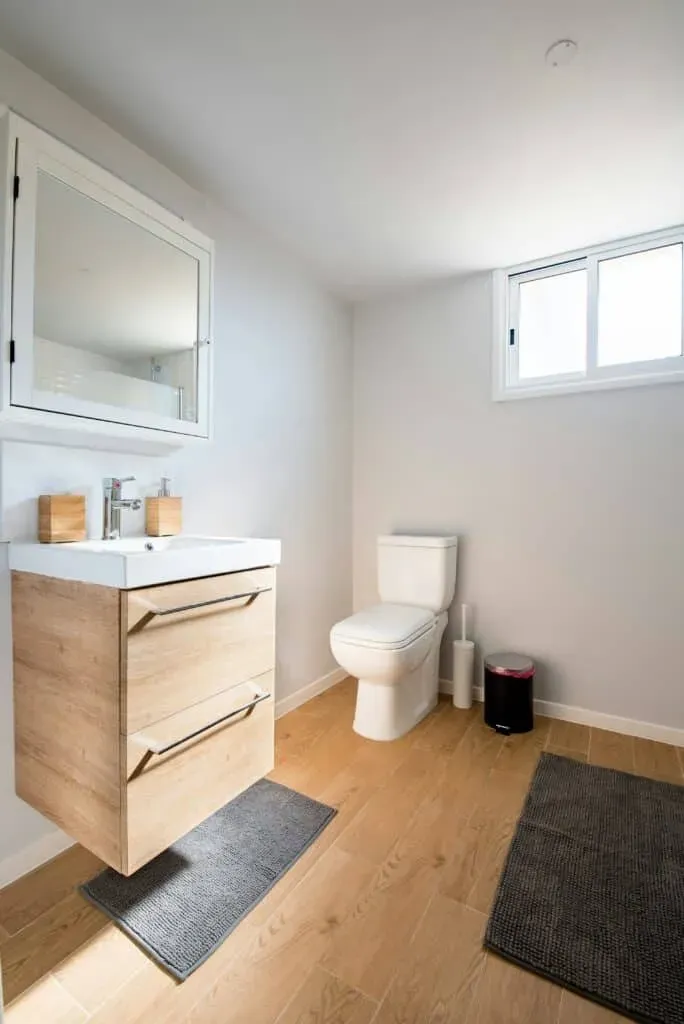 Small residential bathroom with new flooring. Wood-look tiles with two small gray rugs, one in front of the floating vanity.