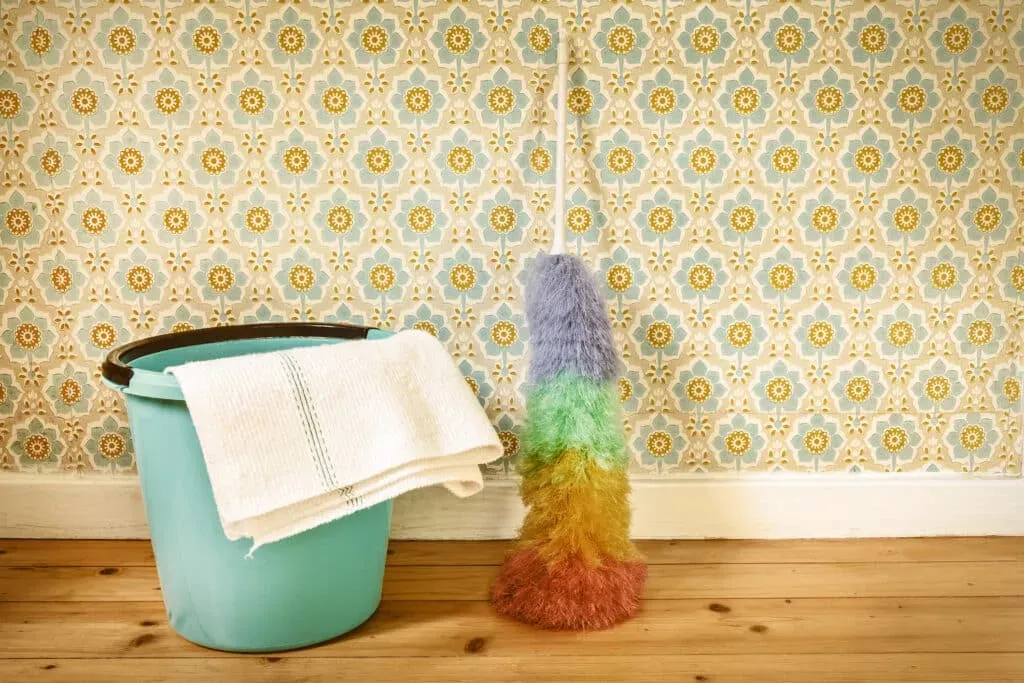 Sepia toned image of a rainbow feather duster on floor, leaning against a wall with flowered wallpaper, next to a teal bucket on a hardwood floor.