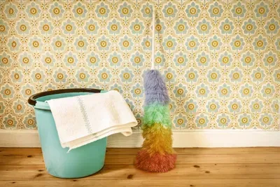 Sepia toned image of a rainbow feather duster on floor, leaning against a wall with flowered wallpaper, next to a teal bucket on a hardwood floor.